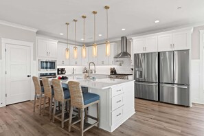 A modern kitchen with white cabinetry, a central island with bar stools, stainless steel appliances, and pendant lighting. - Weekend Rescue: 10BR 10.5BA, Ocean Views, Pool, Hot Tub, Elevator, Dog Friendly, Sleeps 22 (Oak Island)