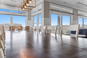 A spacious dining area with a long table and chairs, large windows, and a modern chandelier. - Weekend Rescue: 10BR 10.5BA, Ocean Views, Pool, Hot Tub, Elevator, Dog Friendly, Sleeps 22 (Oak Island)