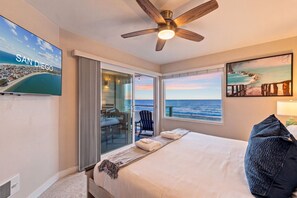 Une chambre d’hôtel équipée d’un lit, d’un ventilateur de plafond, d’une télévision, d’une grande fenêtre donnant sur la plage et d’une porte menant à un balcon.