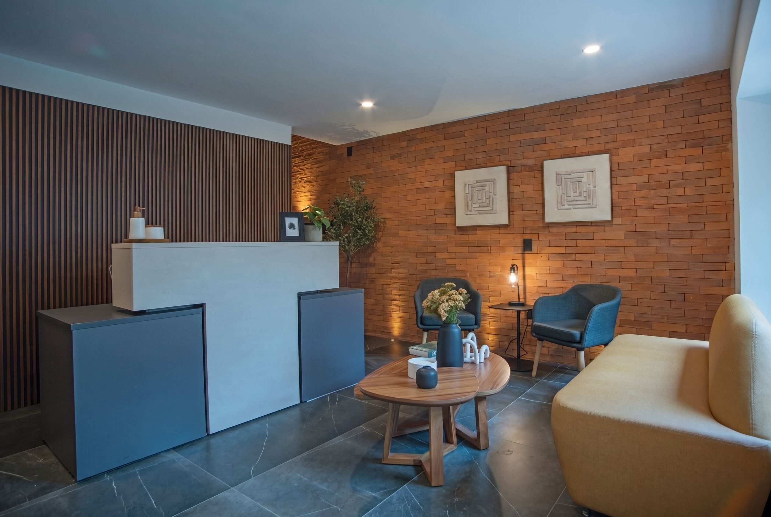 A modern reception area with a brick wall, a reception desk, two chairs, a small table with a vase, and a sofa.