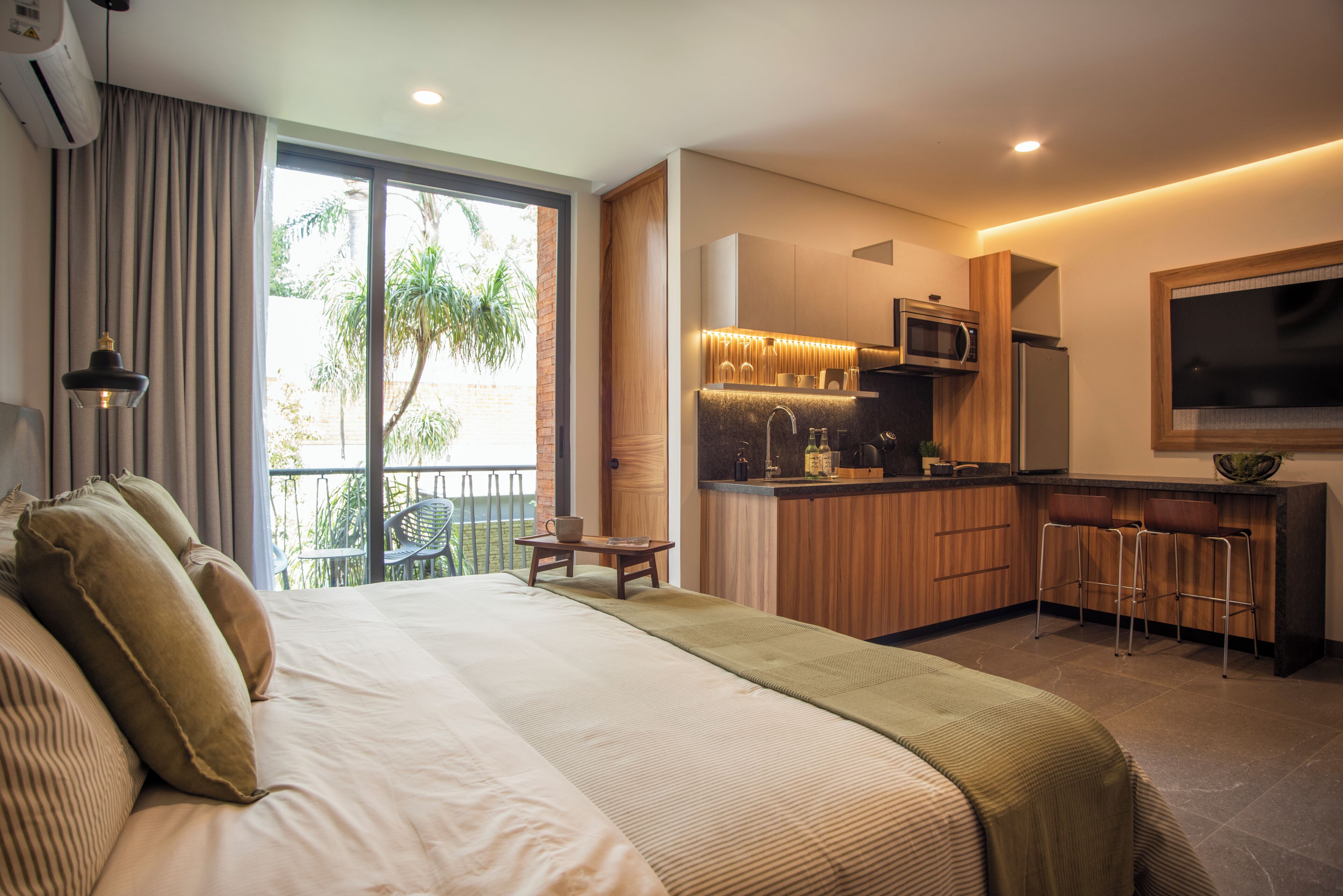 Ein modernes Hotelzimmer mit einem großen Bett, einer Küchenzeile und einem Balkon mit Blick auf Grünflächen.