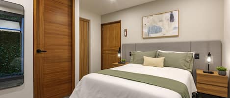 A bedroom with a bed, nightstands, a wall-mounted lamp, a painting, and a window with a view of greenery.