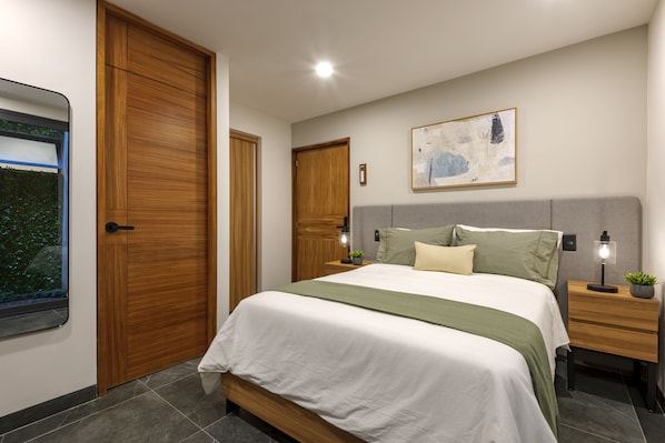 A bedroom with a bed, nightstands, a wall-mounted lamp, a painting, and a window with a view of greenery.