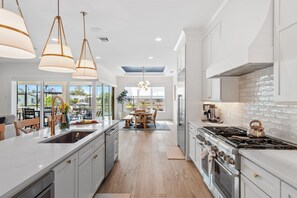 A modern kitchen with a large island, stainless steel appliances, pendant lighting, and a view to the outdoor dining area. - Sunset Riverfront House 4BR | Heated Pool, Hot Tub + Outdoor Kitchen ☀️ (Cocoa Beach)