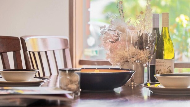 Una mesa de comedor dispuesta con platos, tazones y un florero. Hay dos botellas de vino y copas sobre la mesa.