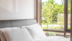 Una cama con sábanas y almohadas blancas, un cabecero tapizado en gris y vistas a la vegetación a través de una ventana.