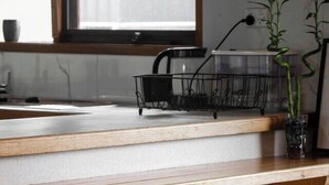 Cocina con encimera de madera, ventana, pileta, cesto de alambre negro, dos taburetes de madera y una planta en maceta.