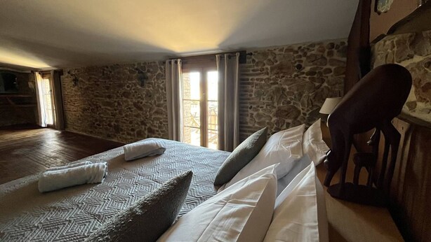 Quarto com parede de pedra, cama com roupa de cama estampada, cabeceira de madeira e vista pela janela.