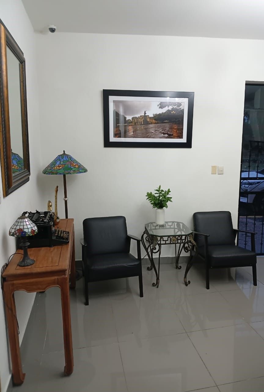 Una habitación con un escritorio de madera, dos sillas negras, una mesita con una planta y un cuadro en la pared.