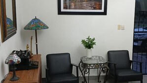Una habitación con un escritorio de madera, dos sillas negras, una mesita con una planta y un cuadro en la pared.