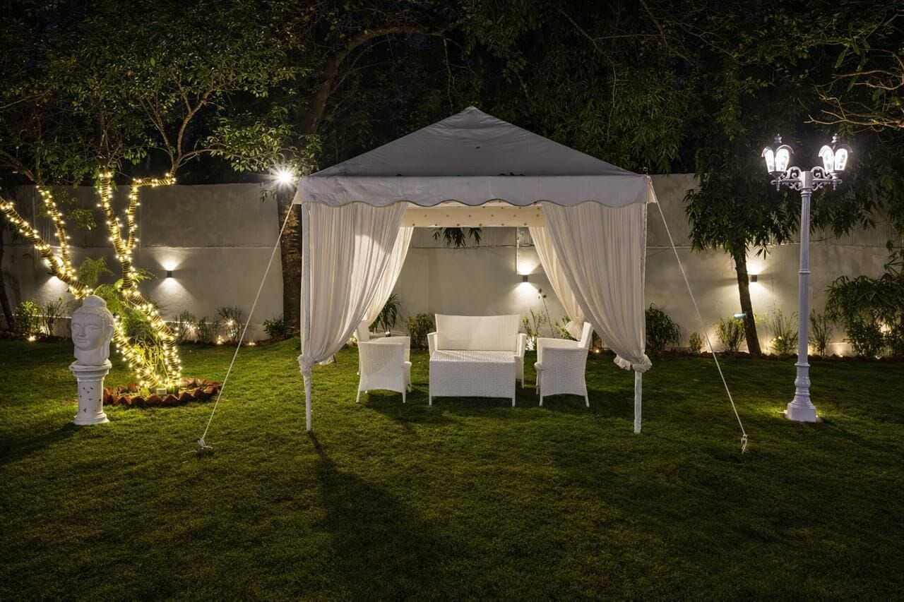 Une tente ornée de tentures blanches, trois chaises blanches et une table, installées sur une pelouse verdoyante, le tout agrémenté de guirlandes lumineuses et d’un lampadaire.