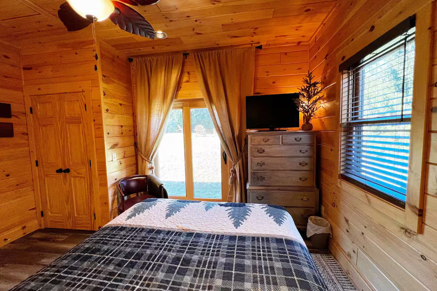 A wooden cabin room with a bed, a dresser, a chair, a television, and a window with blinds.
