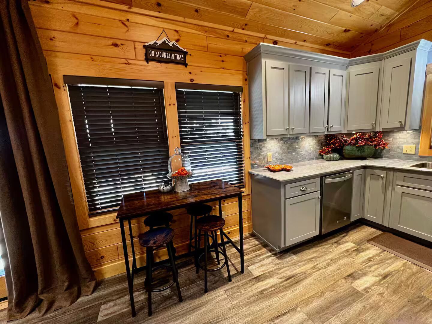Une cuisine avec des murs en bois, des meubles gris, un coin bar avec des tabourets et un panneau indiquant : « HEURE LOCALE DE LA MONTAGNE ».
