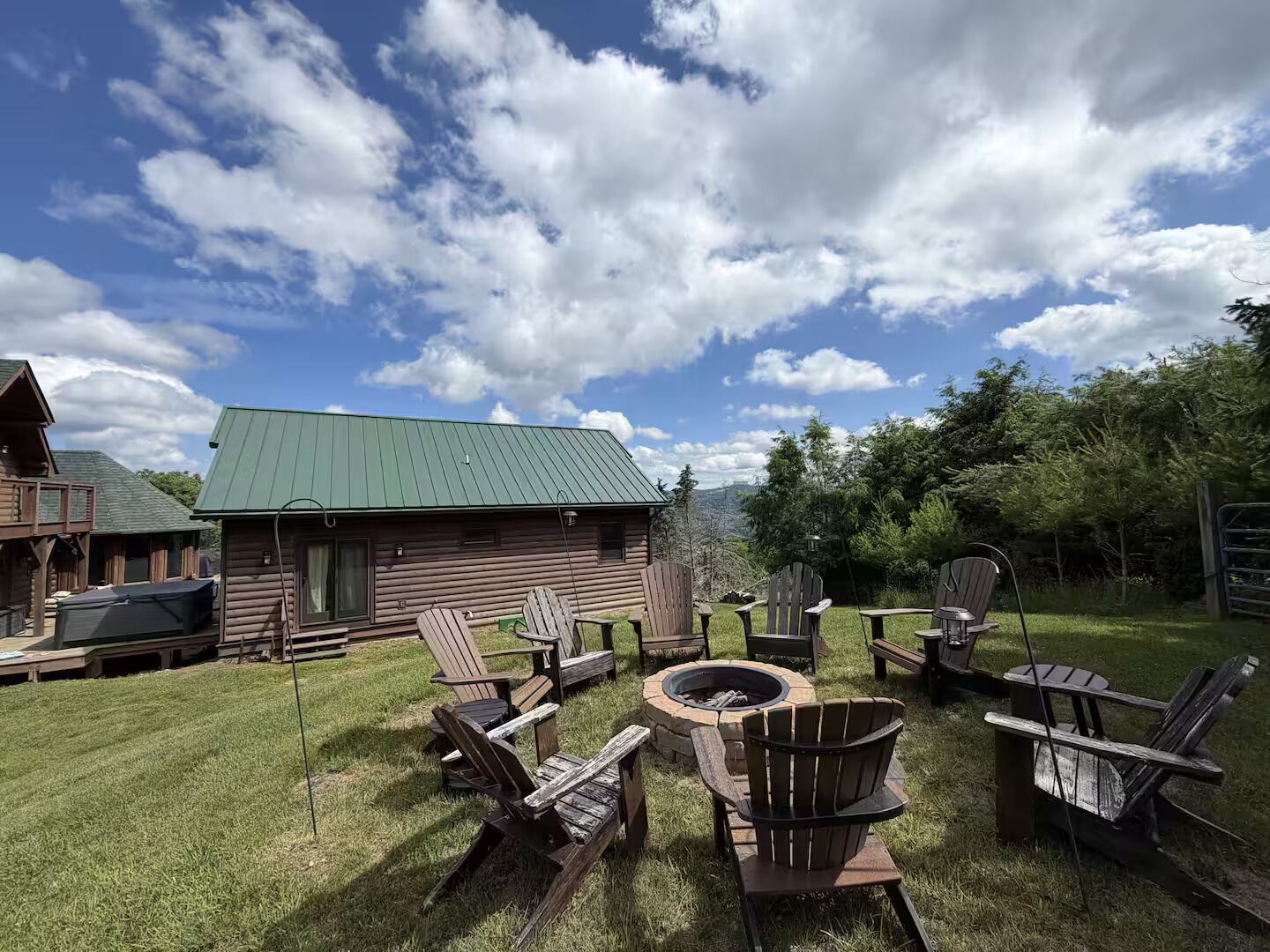 A log cabin with a green roof, surrounded by a grassy yard with wooden Adirondack chairs and a fire pit.