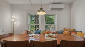 Uma área de jantar com mesa de madeira redonda, cadeiras, luminária pendente e janela com vista para árvores.