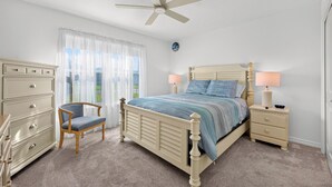 Ein Schlafzimmer mit einem Bett, einem Sessel, einem Schminktisch, einem Nachttisch, einer Lampe und einem Deckenventilator.