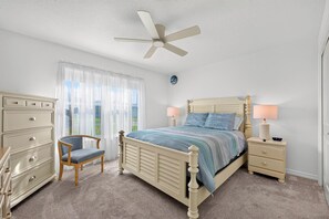 Ein Schlafzimmer mit einem Bett, einem Sessel, einem Schminktisch, einem Nachttisch, einer Lampe und einem Deckenventilator.
