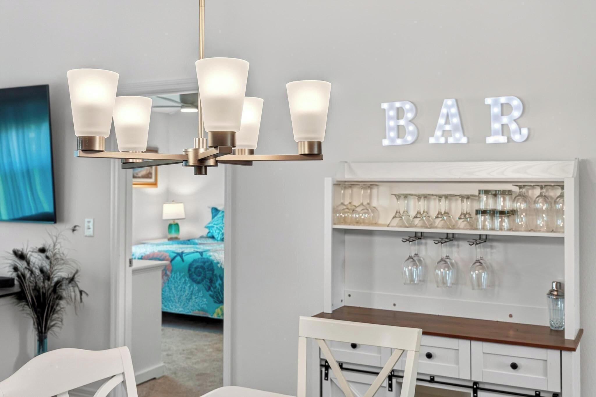 A bar area with a chandelier, a wall-mounted sign reading 'BAR,' and shelves with glasses.