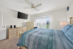 Ein Schlafzimmer mit Bett, Kommode, Fernseher, Deckenventilator und einem Fenster mit Vorhängen.