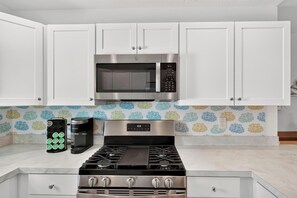 Una cocina con gabinetes blancos, microondas de acero inoxidable, estufa a gas y cafetera.