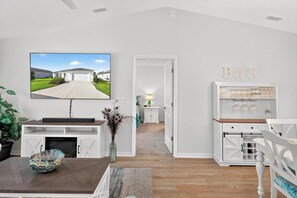 Ein modernes Wohnzimmer mit Flachbildfernseher, Esstisch, Kamin, einer Bar mit Gläserregal und Blick auf ein Haus durch eine Tür.