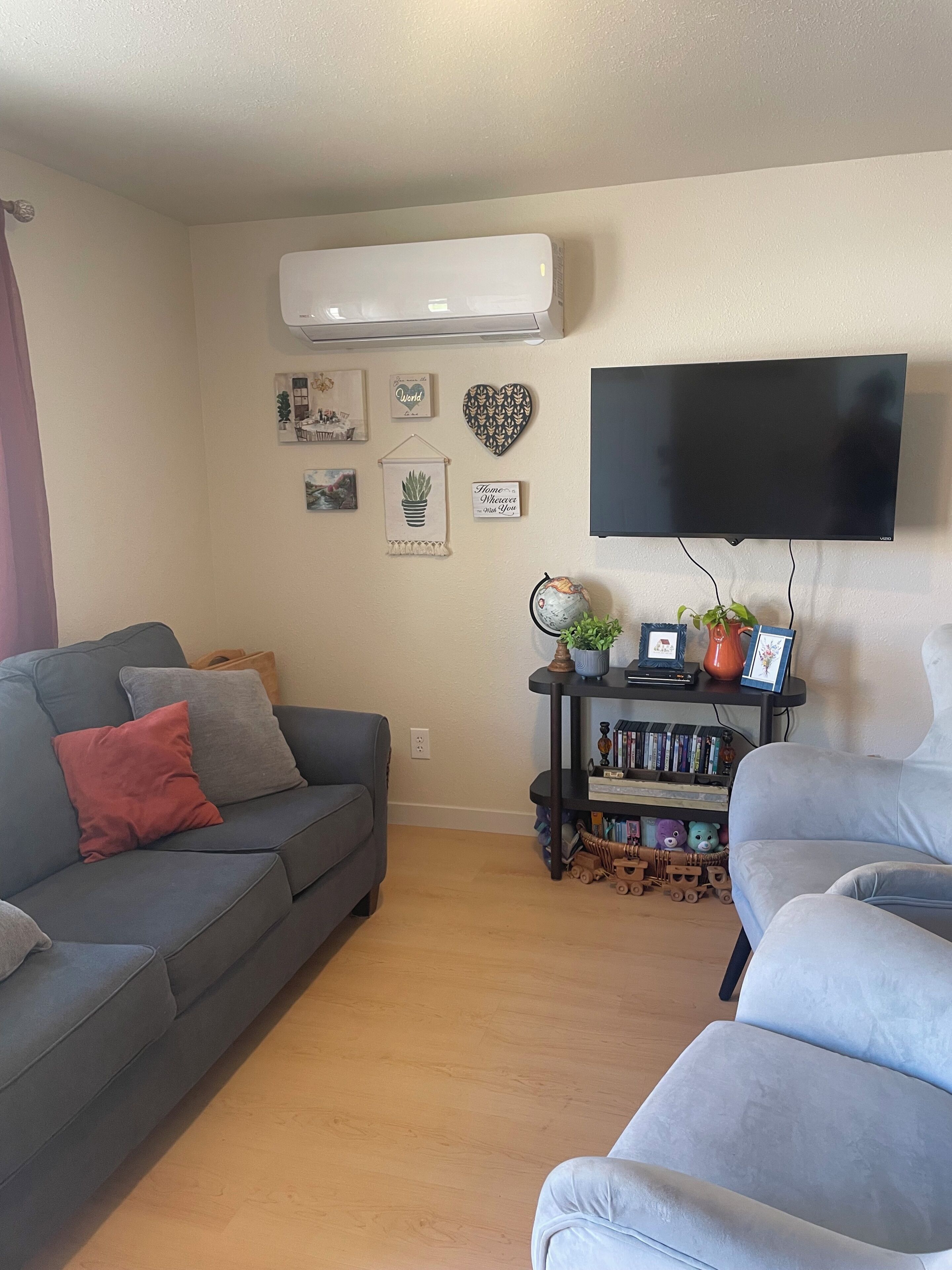 Una sala de estar con un sofá gris, un sillón azul, un televisor de pantalla plana, una estantería y un aire acondicionado de pared.
