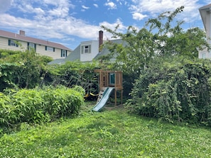 Ein Garten mit einer Holzspielanlage, einer Rutsche und dichtem Grün.