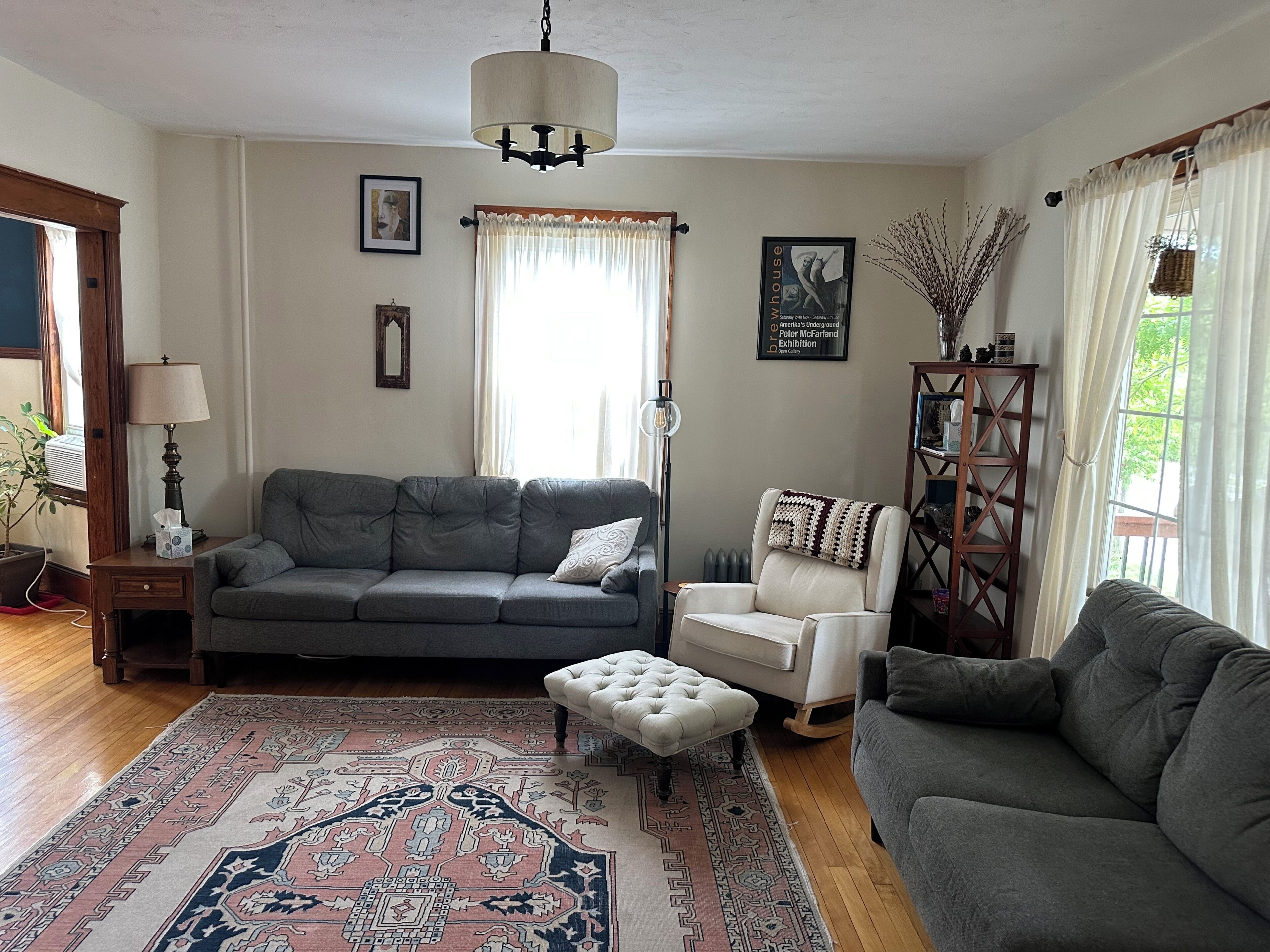 Een woonkamer met een grijze bank, een witte fauteuil, een kamerkleed met dessin, een houten bijzettafeltje en een boekenkast.
