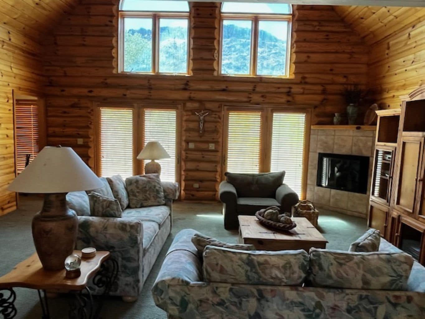 A log cabin interior with a sofa, armchair, lamp, fireplace, and wooden shelves.