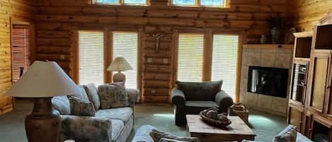 A log cabin interior with a sofa, armchair, lamp, fireplace, and wooden shelves.