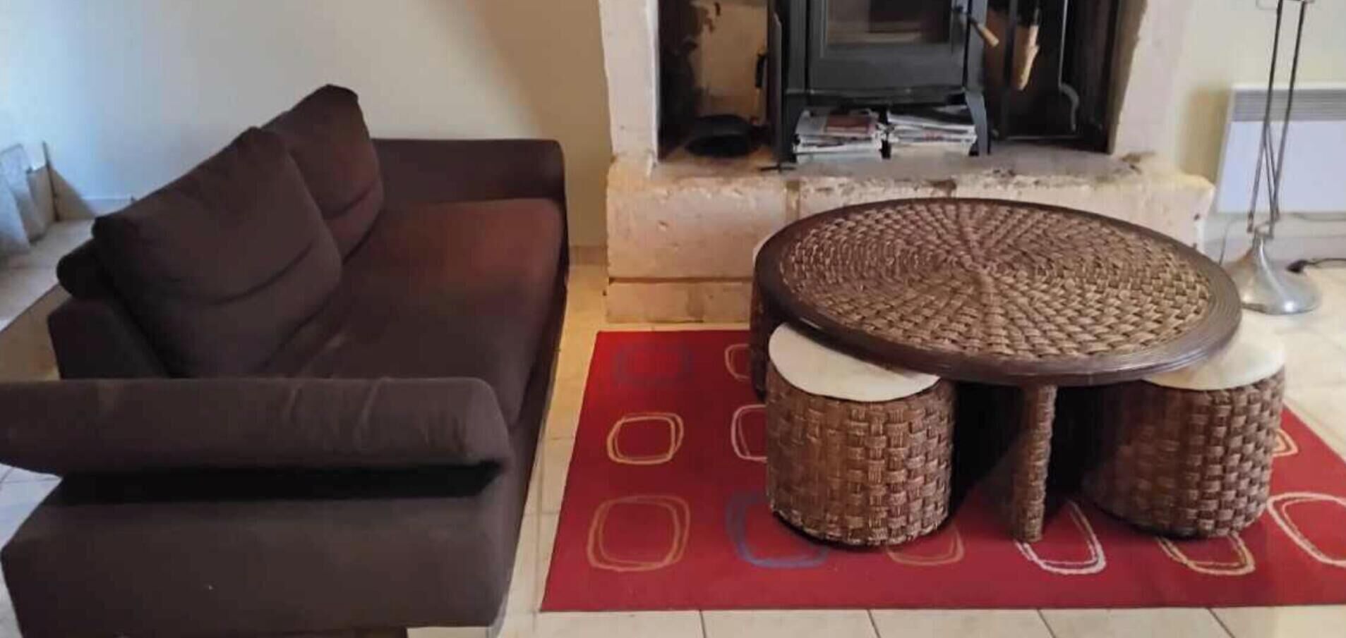 Ein Wohnzimmer mit einem braunen Sofa, einem runden Couchtisch und gewebten Hocker sowie einem roten Teppich mit geometrischen Mustern.