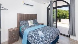 A bedroom with a bed, a nightstand, a drawer unit, an air conditioning unit, and a sliding glass door leading to an outdoor area.