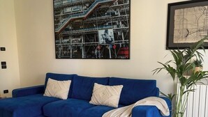 A living room with a blue sectional sofa, a patterned rug, a large abstract painting, and a potted plant. - Welcome to our vacation home, "Azzurro Garda🩵," in Sirmione! (Sirmione)