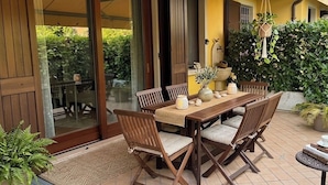 An outdoor patio with a wooden table and chairs, a hanging plant, and a door leading to a garden. - Welcome to our vacation home, "Azzurro Garda🩵," in Sirmione! (Sirmione)