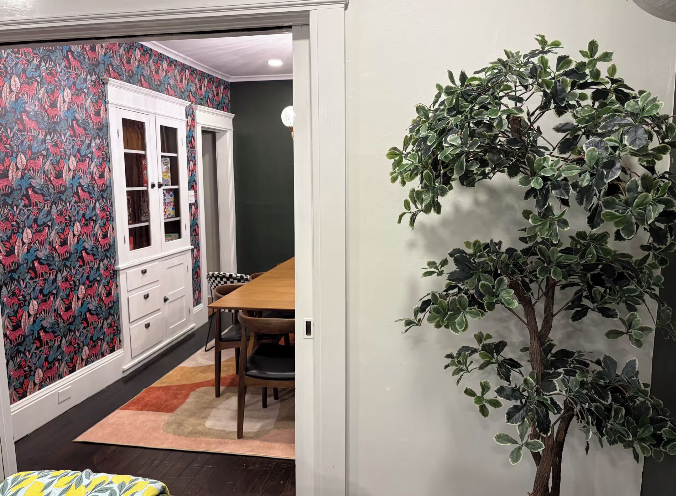 Een kamer met behang met een patroon, een houten eettafel, een kast en een kamerplant.