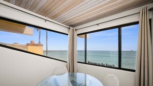 Een kamer met een glazen tafel, beige gordijnen en uitzicht op zee.