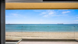 Uitzicht vanaf een balkon op een strand, met een heldere lucht, rustige zee en landschappen in de verte.