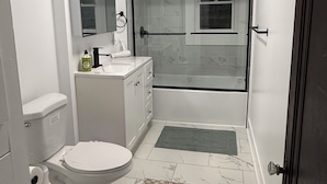 A modern bathroom with a toilet, sink, mirror, shower, and a glass-enclosed shower stall. - Room - 202 - King Bed (Syracuse)