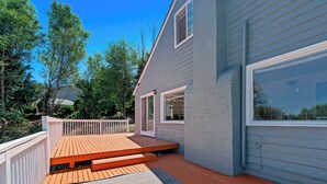 Eine Terrasse mit Holzboden und einer grauen Matte, neben einem Haus mit weißer Geländer und einem großen Fenster.