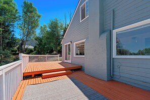 Eine Terrasse mit Holzboden und einer grauen Matte, neben einem Haus mit weißer Geländer und einem großen Fenster.