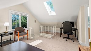 Ein Zimmer mit schrägverdeckter Decke, einem Fenster mit Blick auf Bäume, einem Schreibtisch mit Lampe, einem Stuhl, einem Babybett und einem Gaming-Stuhl.