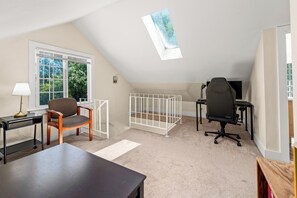 Ein Zimmer mit schrägverdeckter Decke, einem Fenster mit Blick auf Bäume, einem Schreibtisch mit Lampe, einem Stuhl, einem Babybett und einem Gaming-Stuhl.