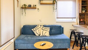 A blue sofa with a remote control on a wooden coffee table, a wooden shelf with plants, and a window with blinds. - Stunning apartment in Stinjan (Stinjan)