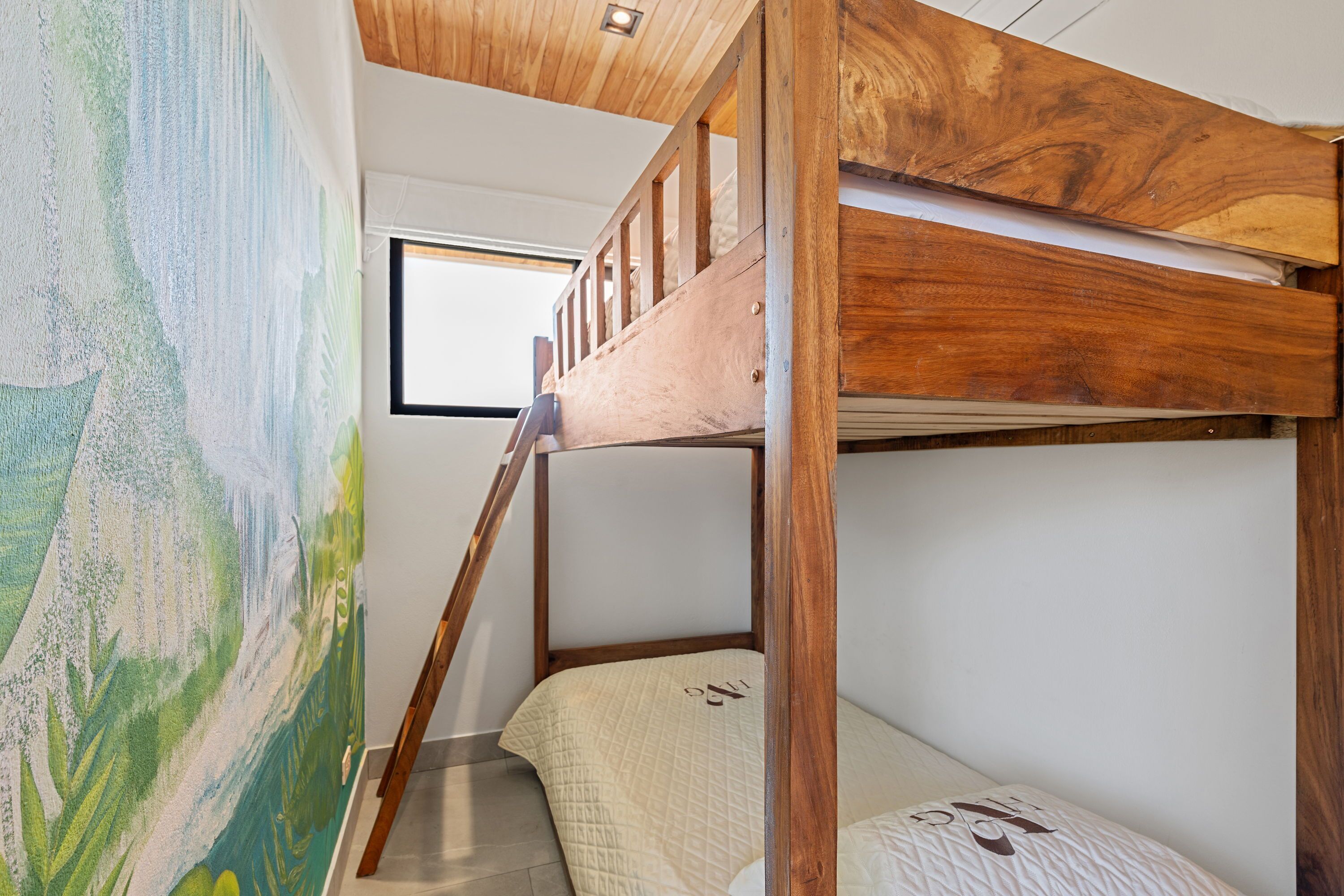 Un lit superposé avec une structure en bois, une fenêtre au-dessus et un tableau sur le mur adjacent.