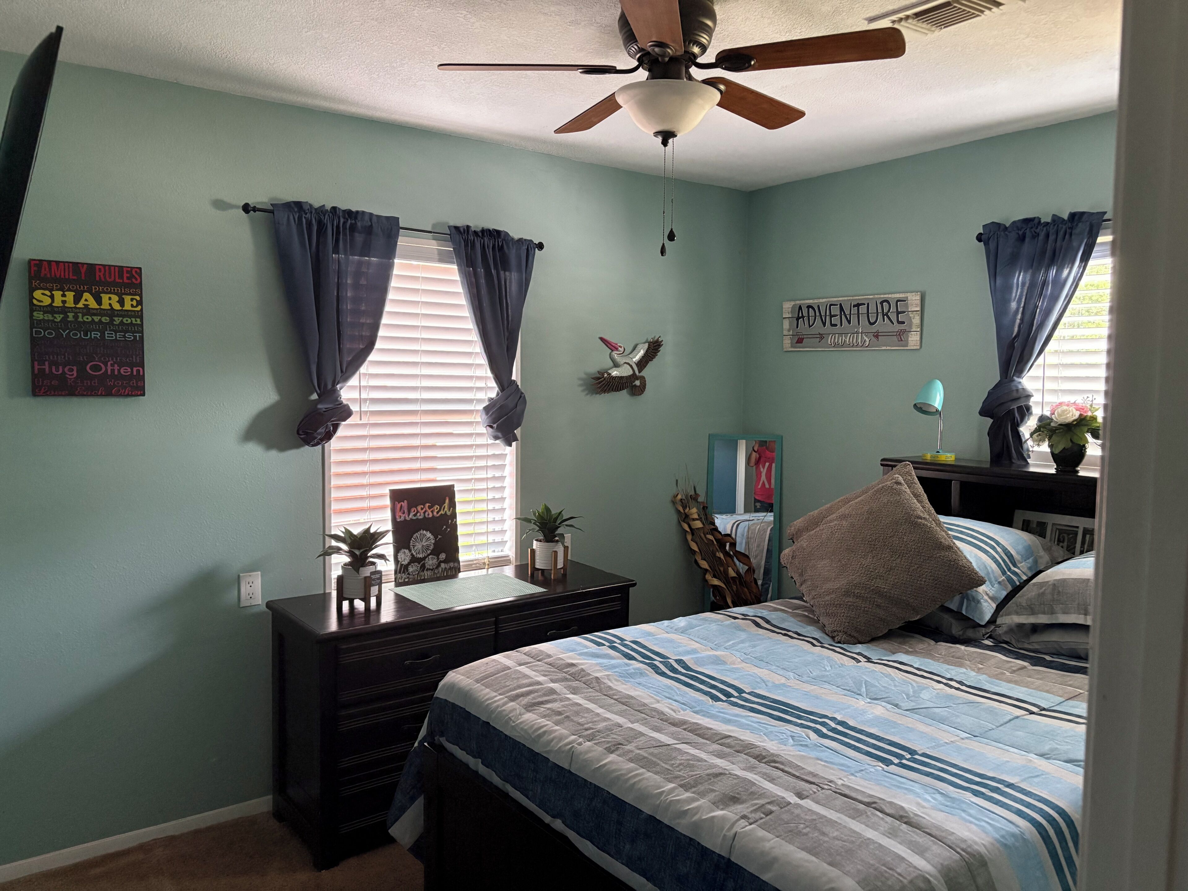 Une chambre à coucher avec un lit, une commode, un ventilateur de plafond et une fenêtre avec des rideaux.