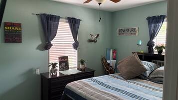 Une chambre à coucher avec un lit, une commode, un ventilateur de plafond et une fenêtre avec des rideaux.