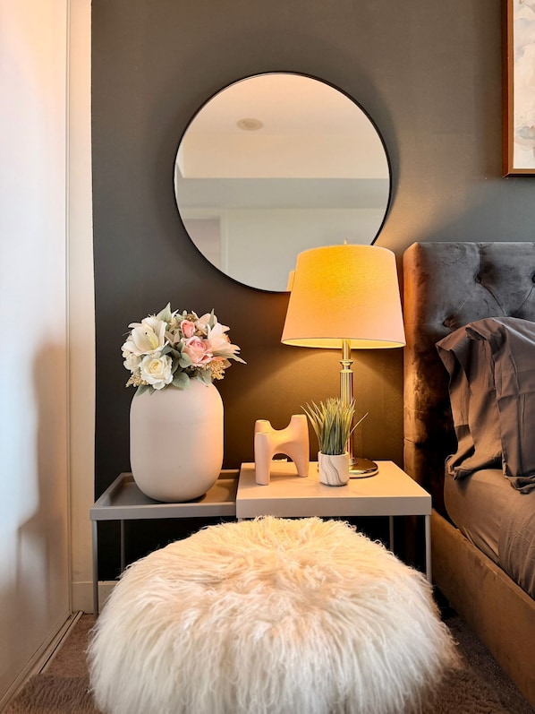 Una habitación con un espejo redondo, una lámpara de noche, un otomano blanco y mullido, un jarrón con flores y un cabecero capitonado.