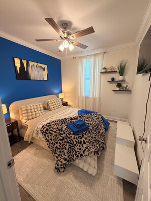Ein Schlafzimmer mit einem Bett, einem Deckenventilator, einem Nachttisch, einer Lampe, einem Fenster mit Vorhängen und einem an der Wand befestigten Regal mit Pflanzen.