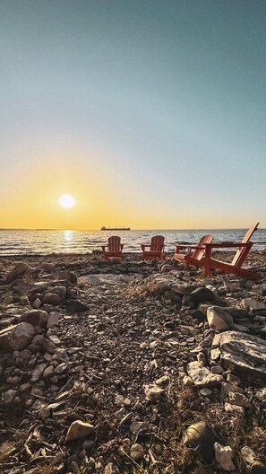 A rocky beach with three wooden chairs facing the ocean at sunset.