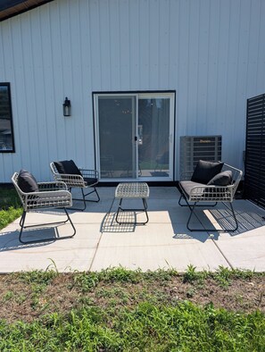 A patio with two metal-framed chairs and a small table, a sliding glass door, and a large air conditioning unit. - Dune Haus + Indiana National Dunes Park (Portage)